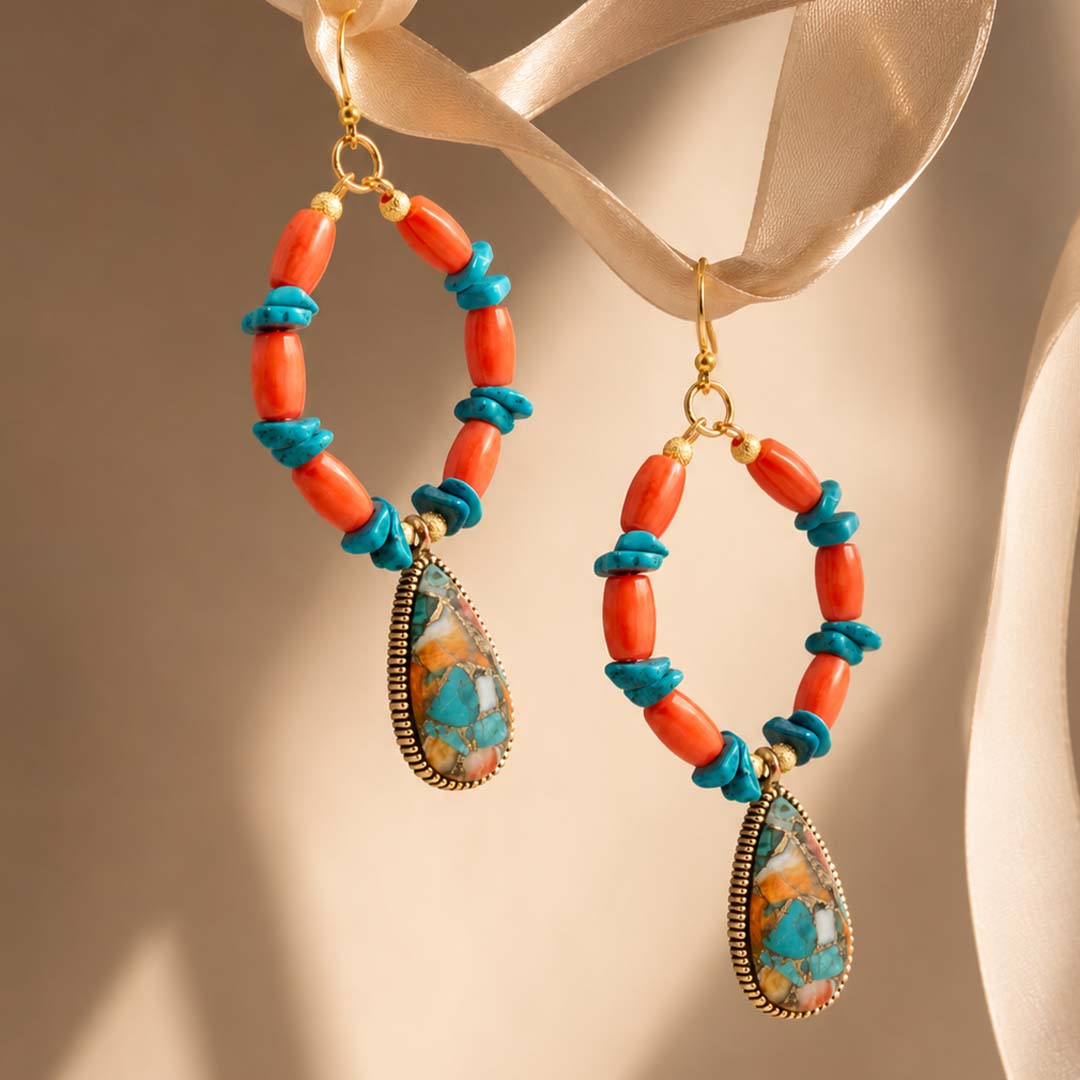 A pair of bohemian-style drop earrings with coral-orange beads, turquoise-blue wrapped accents, and teardrop-shaped marbled turquoise and gold pendants, hanging from a beige satin ribbon against a warm beige background.