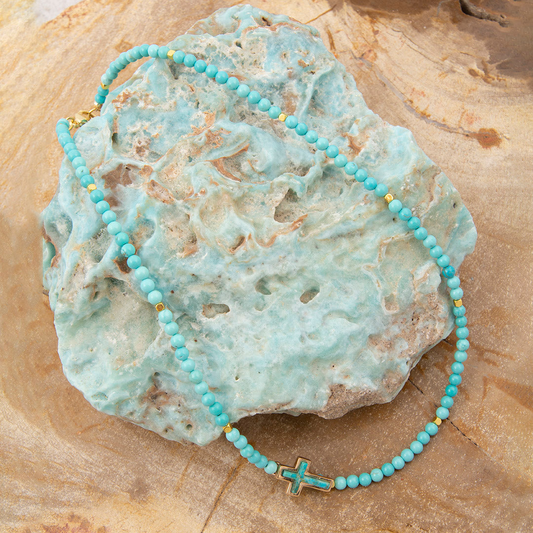 Turquoise beaded necklace with a gold cross pendant on a textured stone surface