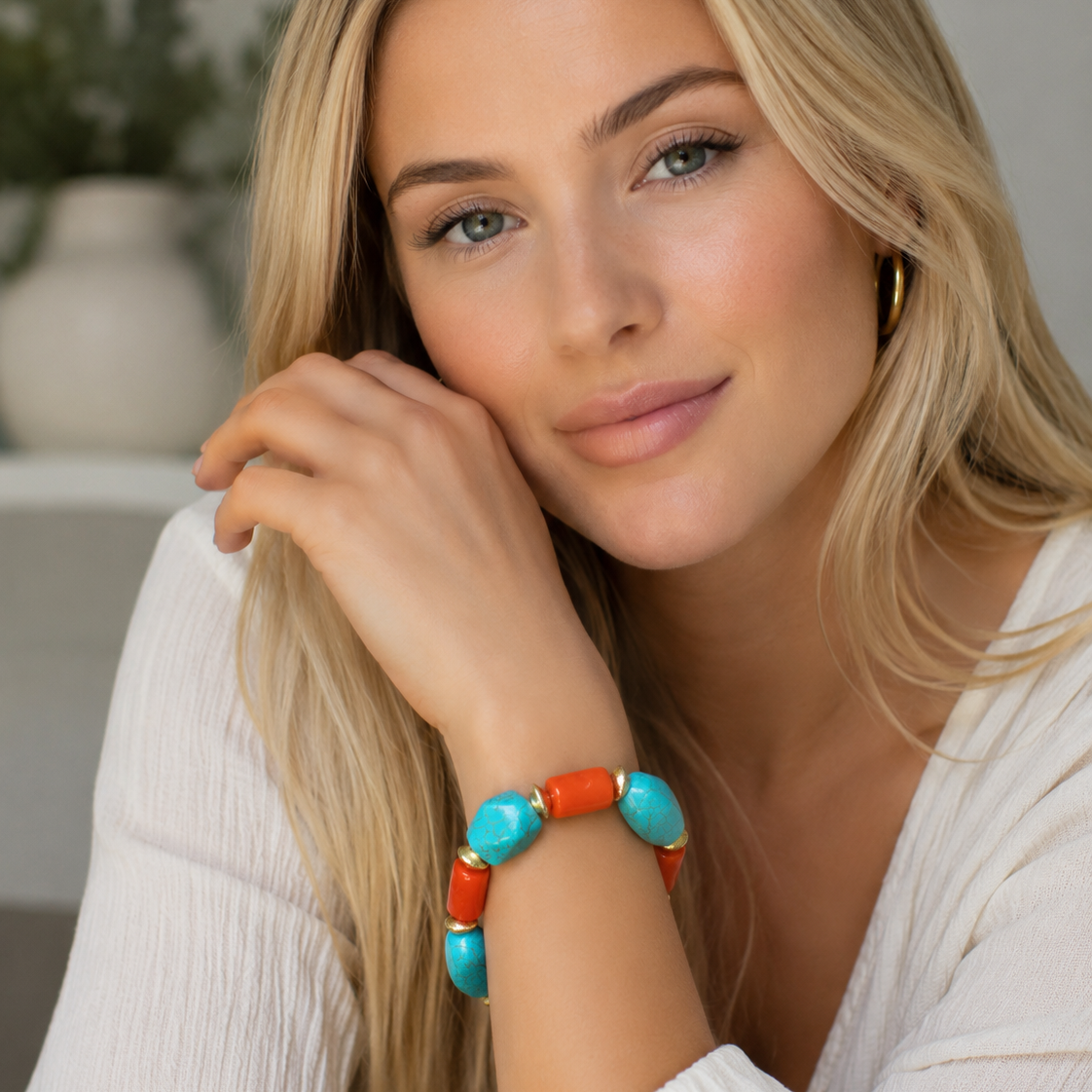 A blonde woman wearing a chunky beaded bracelet with turquoise and orange-red beads separated by gold spacer rings, resting her chin on her hand in a bright indoor setting.