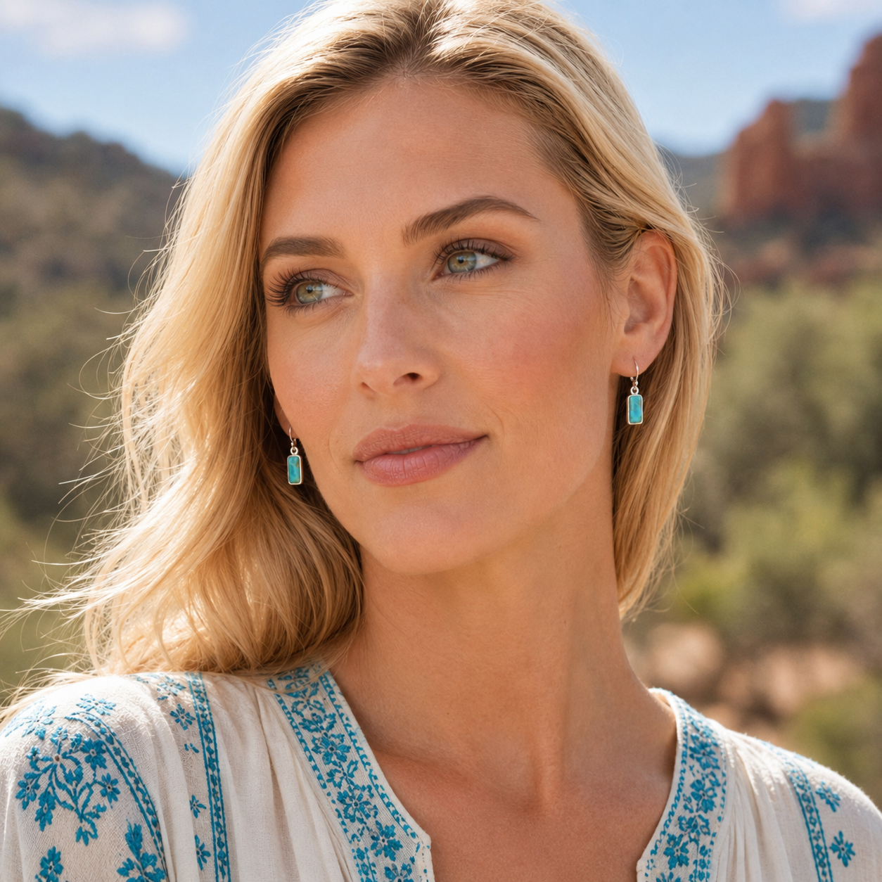 A close-up of a woman with blonde hair wearing small turquoise drop earrings and a cream blouse with blue embroidery, set against a blurred outdoor desert landscape.