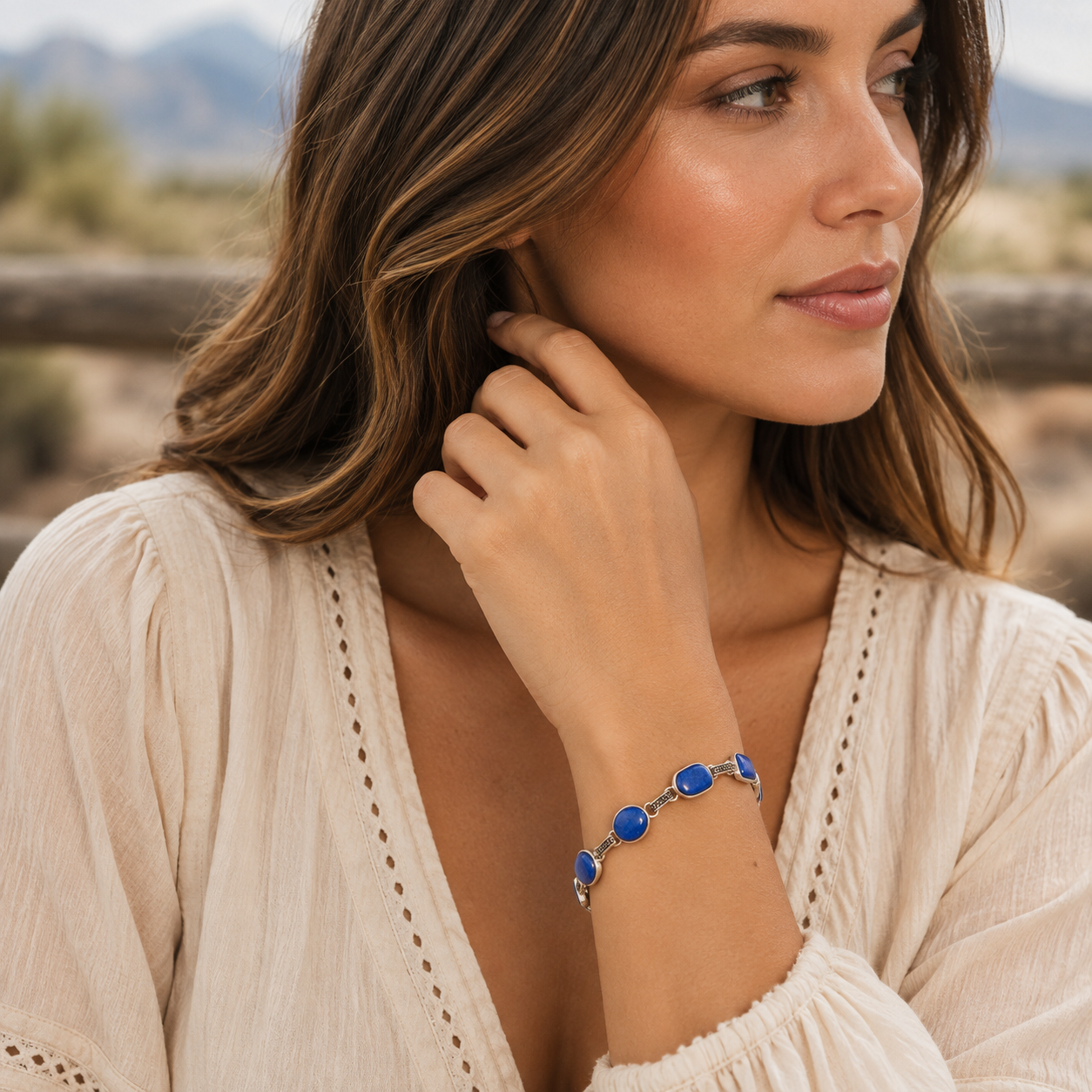 A woman wearing a silver-toned bracelet with three round blue gemstones, shown in a close-up outdoor portrait. The background is a softly blurred desert landscape.