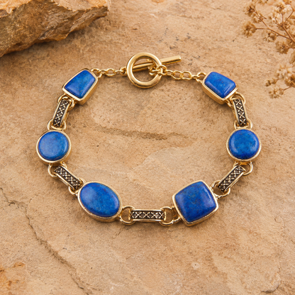 A gold-tone bracelet with six oval and rectangular blue lapis lazuli stones, displayed on a textured tan stone surface.