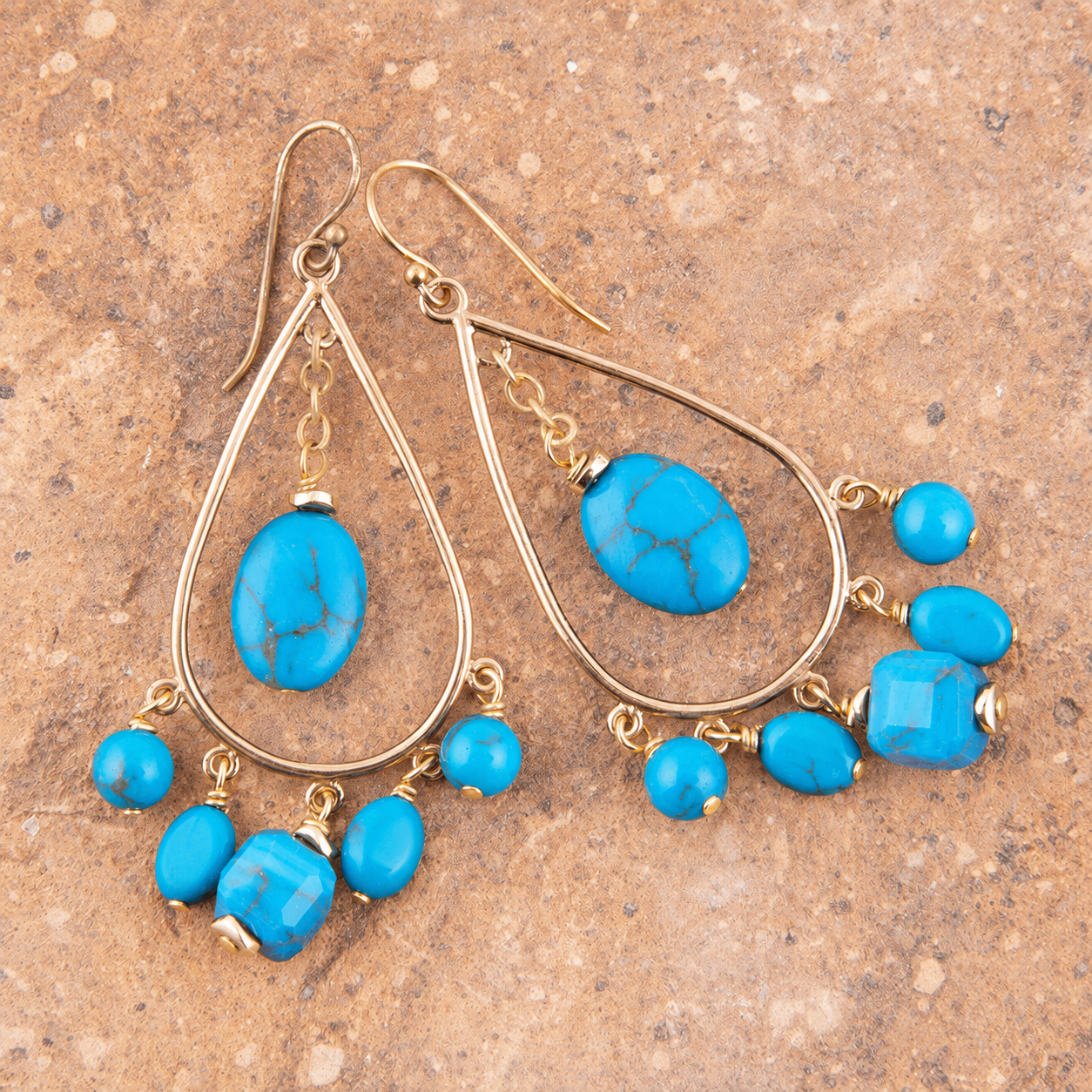 A pair of gold-tone teardrop dangle earrings with turquoise blue marbled beads and small round beads, displayed on a textured brown stone surface.