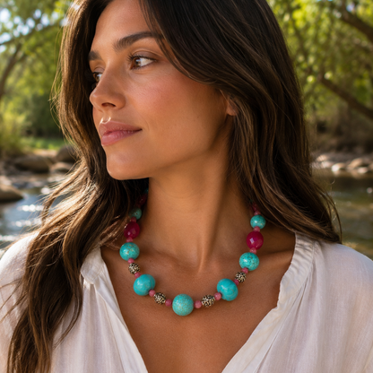 A woman wearing a chunky beaded necklace with large turquoise-blue beads, bright pink beads, and small metallic spacer beads, photographed outdoors near water with trees in the background.