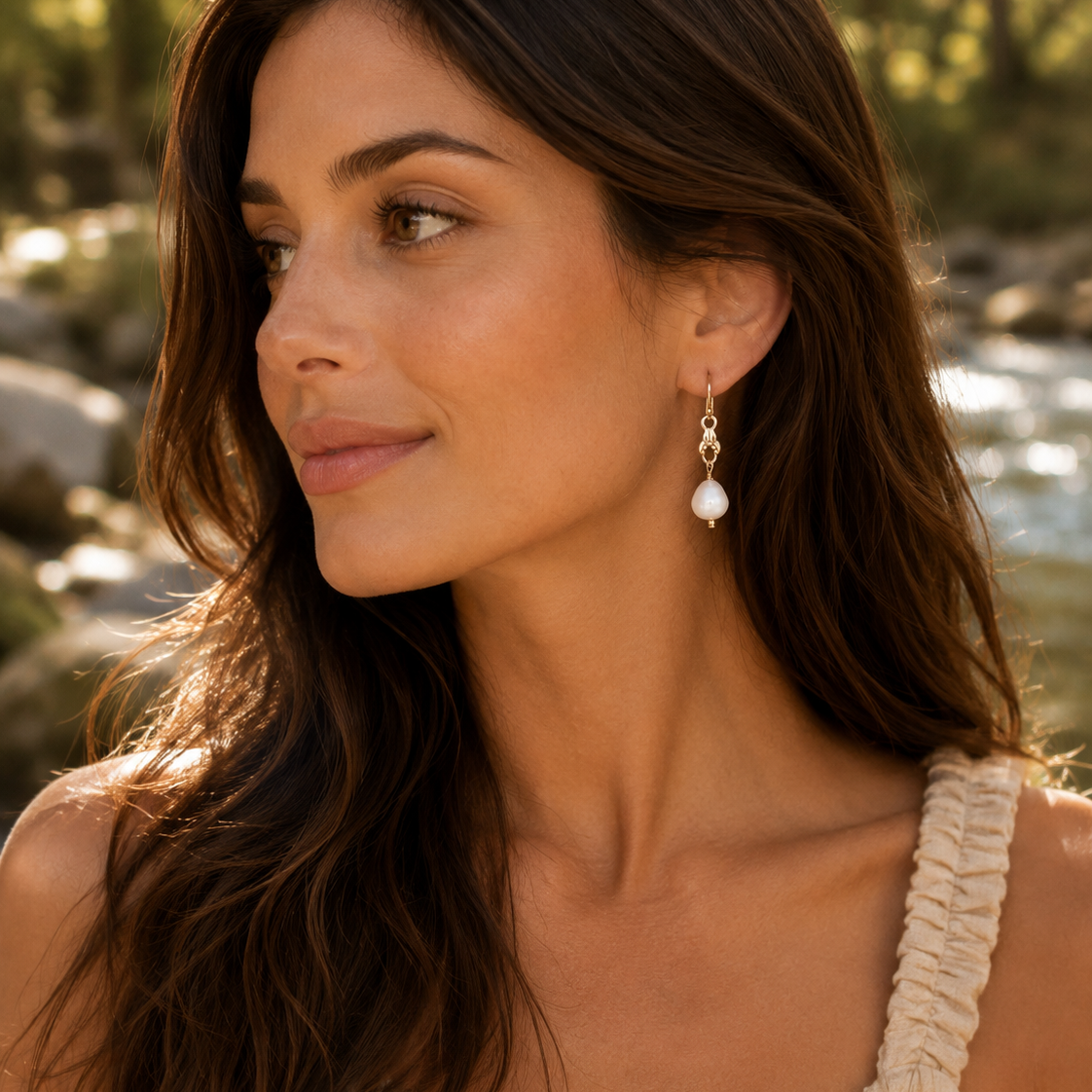 Woman wearing pearl earrings in a natural setting with water and trees.