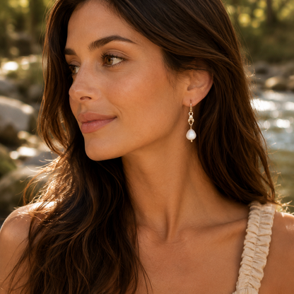 Woman wearing pearl earrings in a natural setting with water and trees.