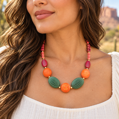 A close-up of a woman wearing a colorful beaded necklace with green, orange, and pink beads, photographed outdoors with a blurred natural background.