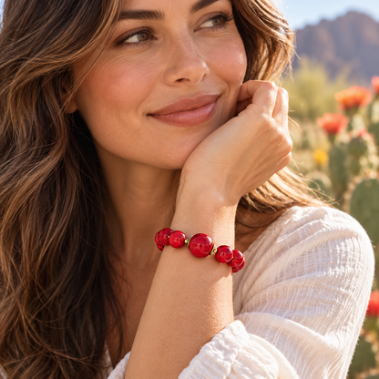 A woman wearing a red beaded bracelet with gold spacer beads, posing outdoors in a desert setting with cacti and mountains in the background.