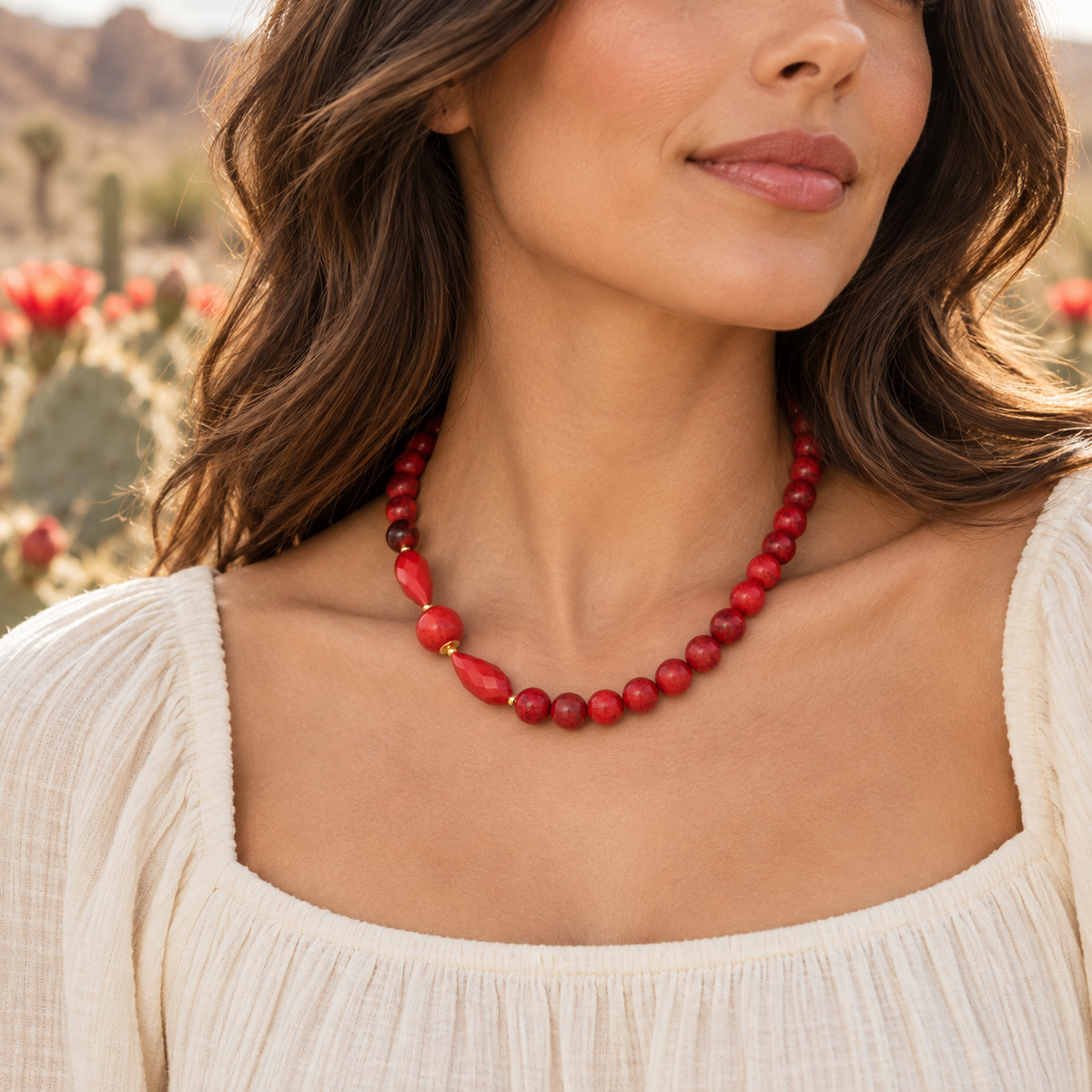 A close-up of a woman wearing a red beaded necklace with a chunky red pendant and small gold spacer beads, paired with a cream-colored top, set against a blurred desert landscape background.
