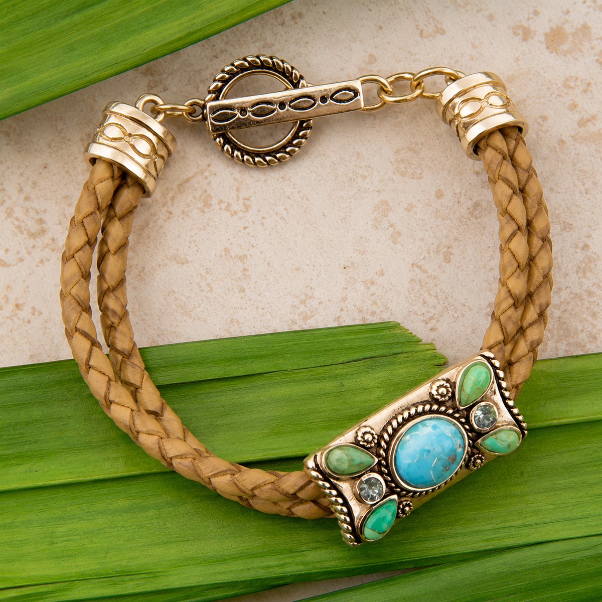 A braided tan leather bracelet with ornate gold-tone metal hardware, featuring a large turquoise and green stone clasp, displayed on green leaves over a light beige textured surface.