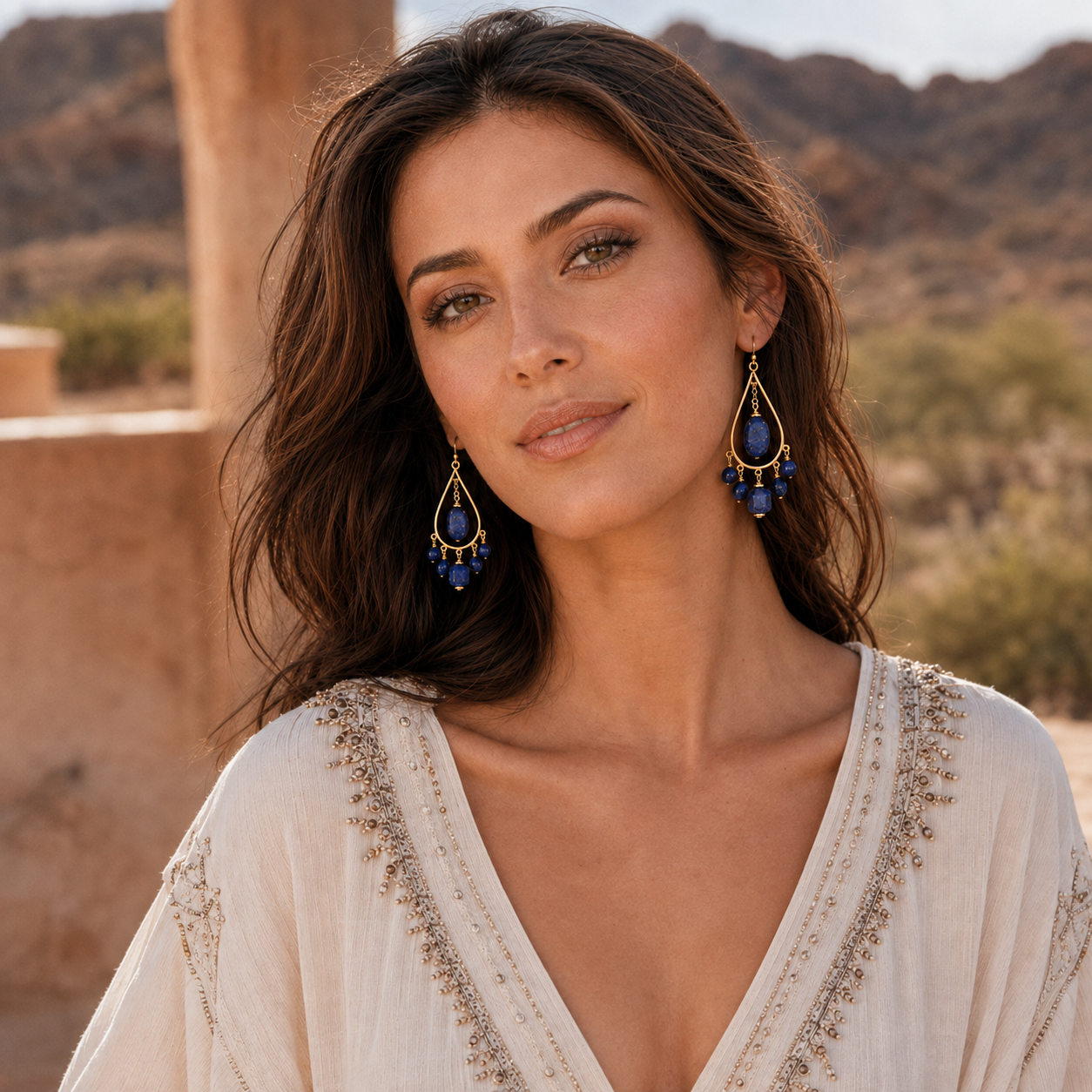 A woman wearing large gold and blue teardrop dangle earrings, photographed outdoors against a warm desert landscape with tan stone structures.