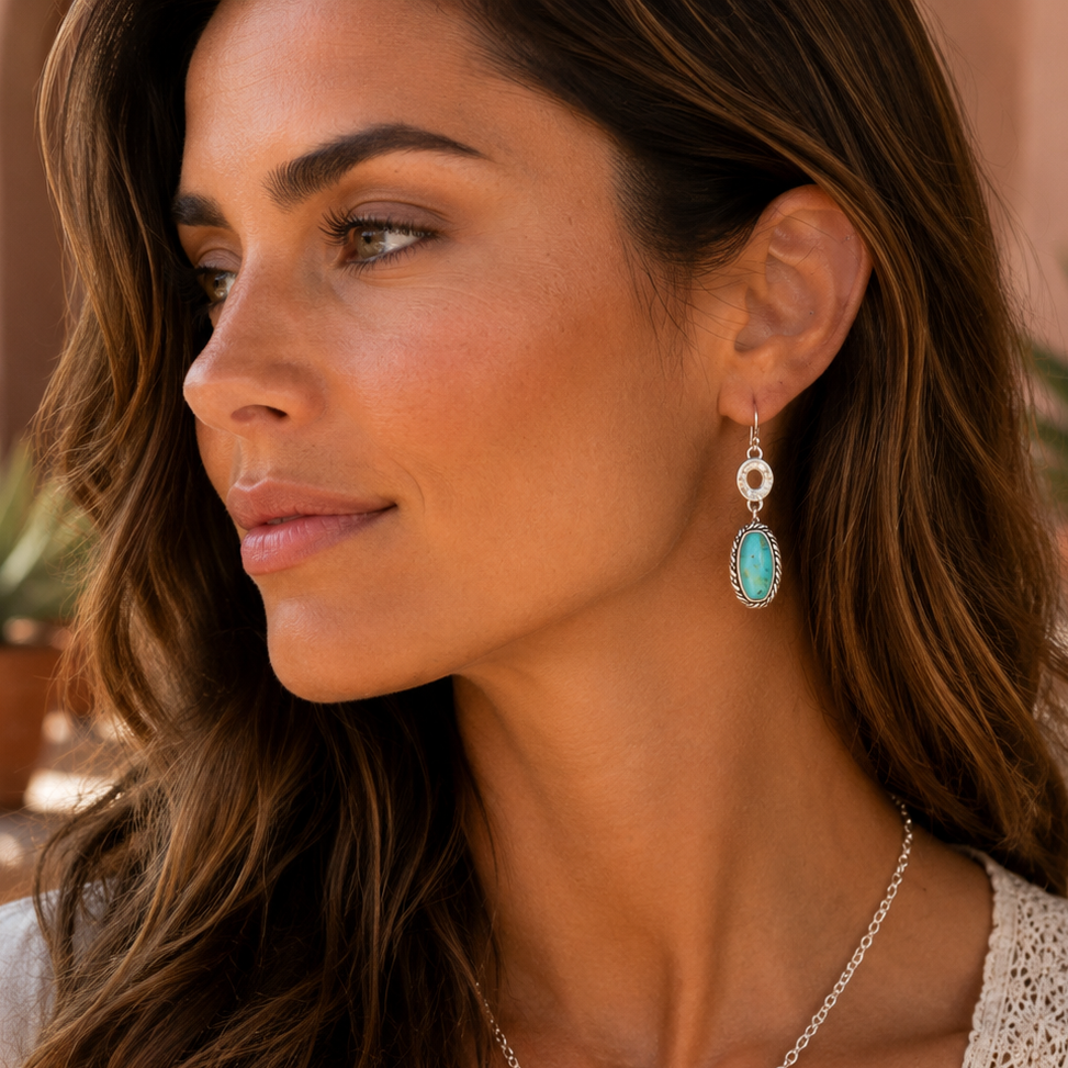 A woman wearing a silver-tone necklace and matching drop earrings with oval turquoise-blue stones, shown in a warm indoor setting.