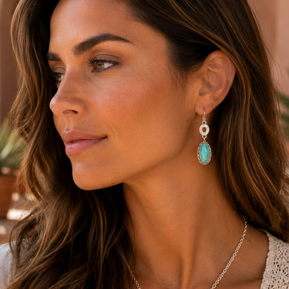A woman wearing a silver-tone necklace and matching drop earrings with oval turquoise-blue stones, shown in a warm indoor setting.