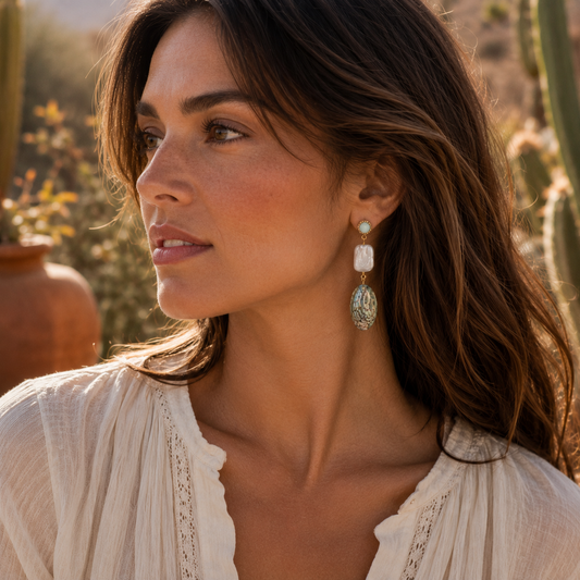 A woman in a white blouse wearing a dangling earring with a white rectangular stone, a light blue round stone, and an oval light blue-green stone with silver-toned detailing, photographed outdoors in warm natural light.