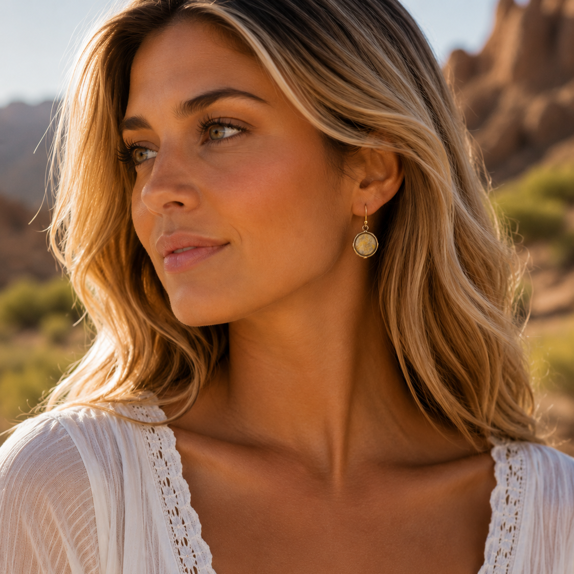 A woman with blonde-highlighted hair wearing a white lace-trim top and a small gold dangle earring, photographed outdoors against a blurred rocky desert background.