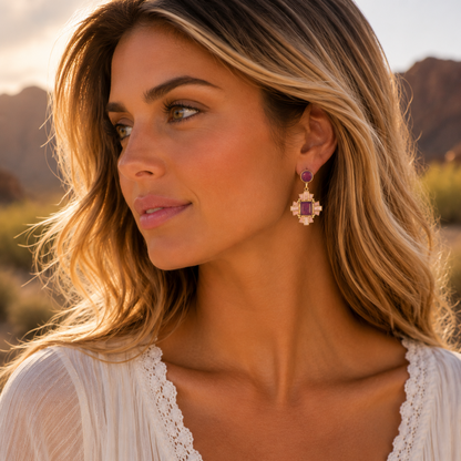 A woman in a white lace-trim top wearing a dangling cross-shaped earring with a purple stone and gold-toned details, photographed outdoors in warm golden hour light.