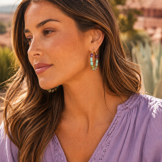 A woman in a lavender eyelet top wearing a gold drop earring with turquoise and purple beads, photographed in warm outdoor light.
