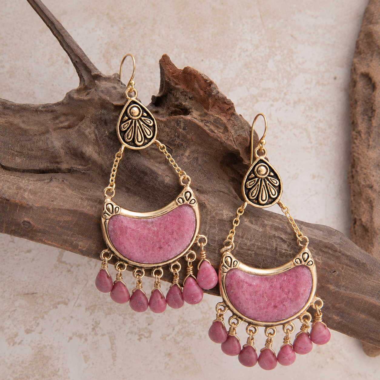 A pair of bohemian gold-toned dangle earrings with pink stone inlays and small pink teardrop beads, displayed on a piece of driftwood against a light textured background.