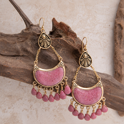 A pair of bohemian gold-toned dangle earrings with pink stone inlays and small pink teardrop beads, displayed on a piece of driftwood against a light textured background.