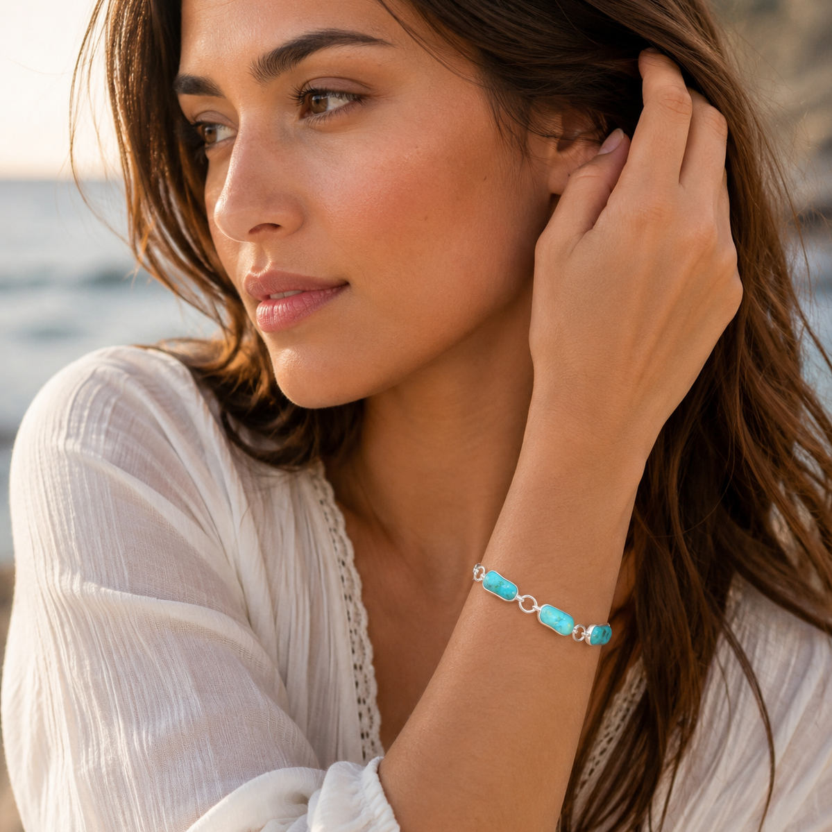 A woman wearing a delicate bracelet with turquoise-colored oval beads and silver-toned links, posing near a beach with her hand on her ear.