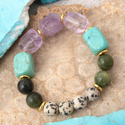A beaded stretch bracelet featuring faceted turquoise, purple amethyst, green jasper, black tourmaline, and spotted dogtooth beads separated by small gold spacer rings, displayed on a textured beige crystal surface with turquoise rock edges.