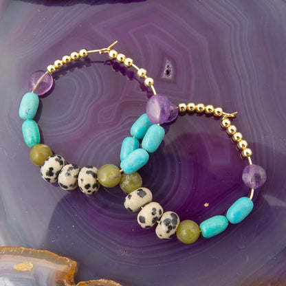 Two beaded hoop earrings displayed on a purple agate slice. The hoops feature gold-tone beads, light blue turquoise beads, purple faceted beads, olive green beads, and spotted white and black labradorite beads.