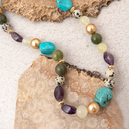 A beaded strand necklace with turquoise, amethyst, olive green, white, and gold beads, styled on a textured beige surface with mineral slices.