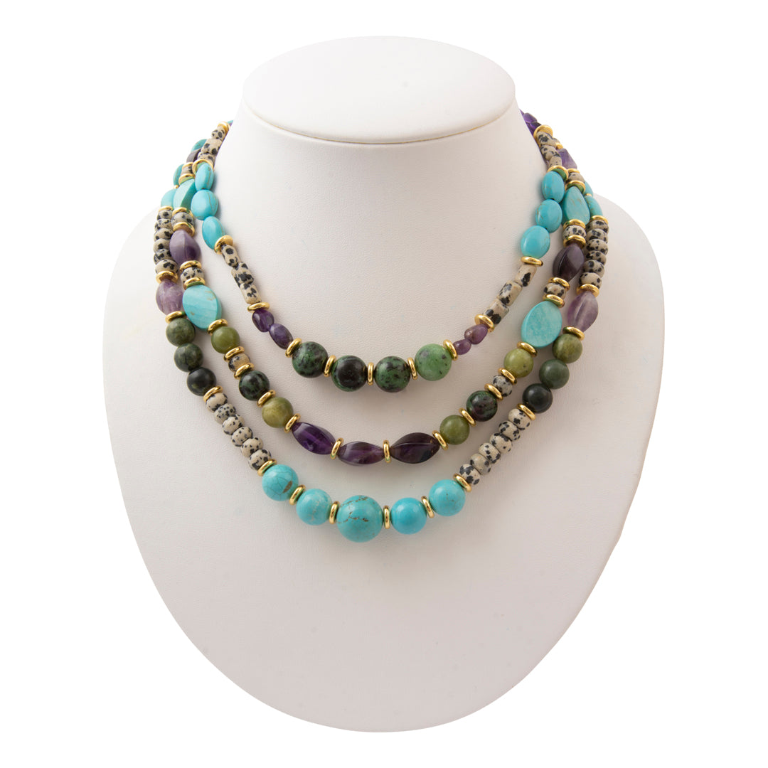 A three-strand beaded necklace displayed on a white jewelry bust. The necklace features turquoise, purple amethyst, olive green, and dark green beads, accented with gold spacers and textured silver beads.
