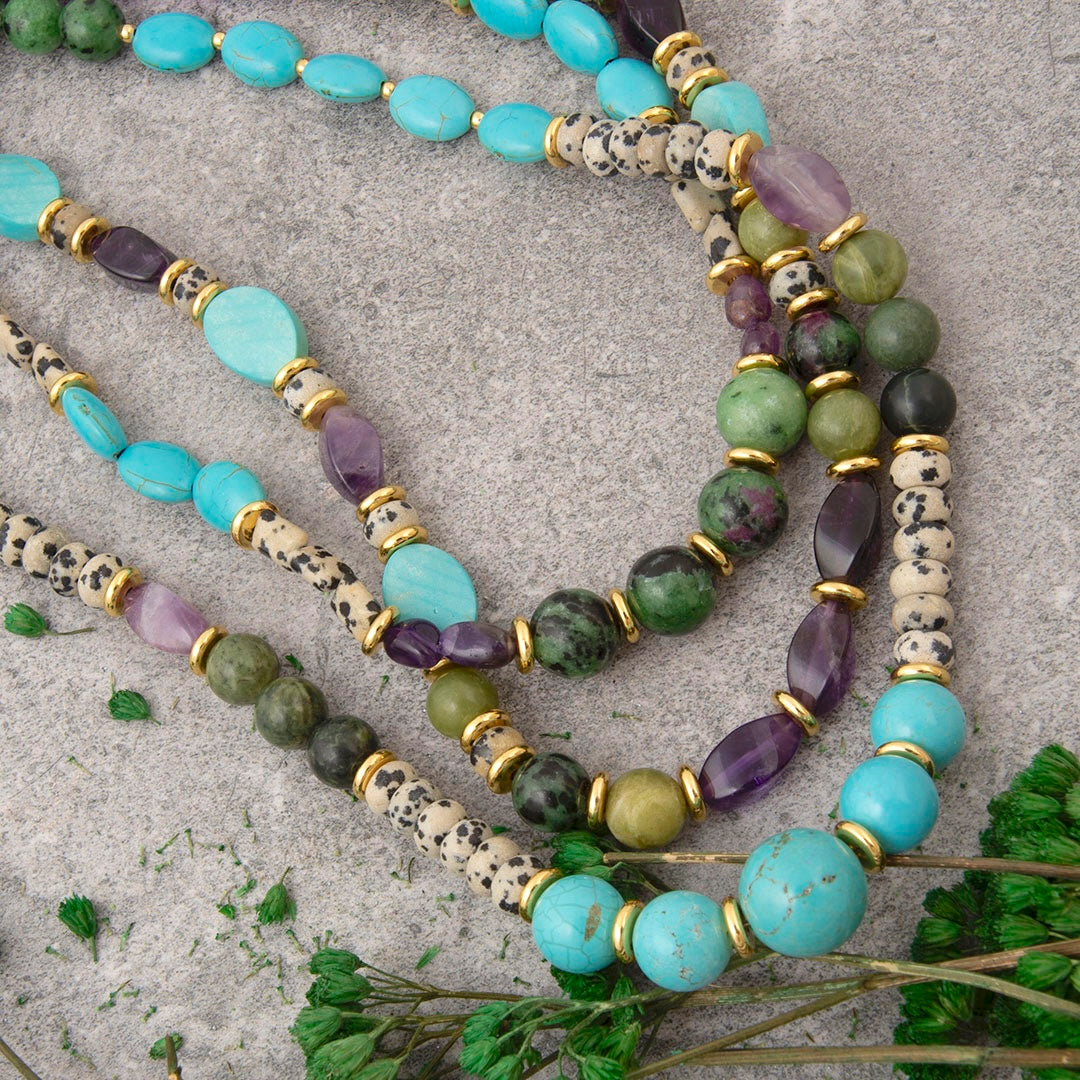 A layered strand of beaded jewelry laid flat on a textured gray surface, featuring turquoise, light blue, green, and purple stones with gold spacer rings and cream-and-black speckled cylindrical beads. Small green leaves are placed near the bottom.