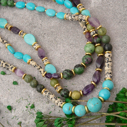 A layered strand of beaded jewelry laid flat on a textured gray surface, featuring turquoise, light blue, green, and purple stones with gold spacer rings and cream-and-black speckled cylindrical beads. Small green leaves are placed near the bottom.