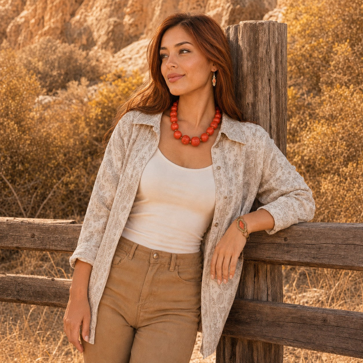 A woman leans against a rustic wooden fence in a desert landscape, wearing a light beige patterned button-up shirt over a white tank top and tan pants, accessorized with a chunky orange-red beaded necklace.