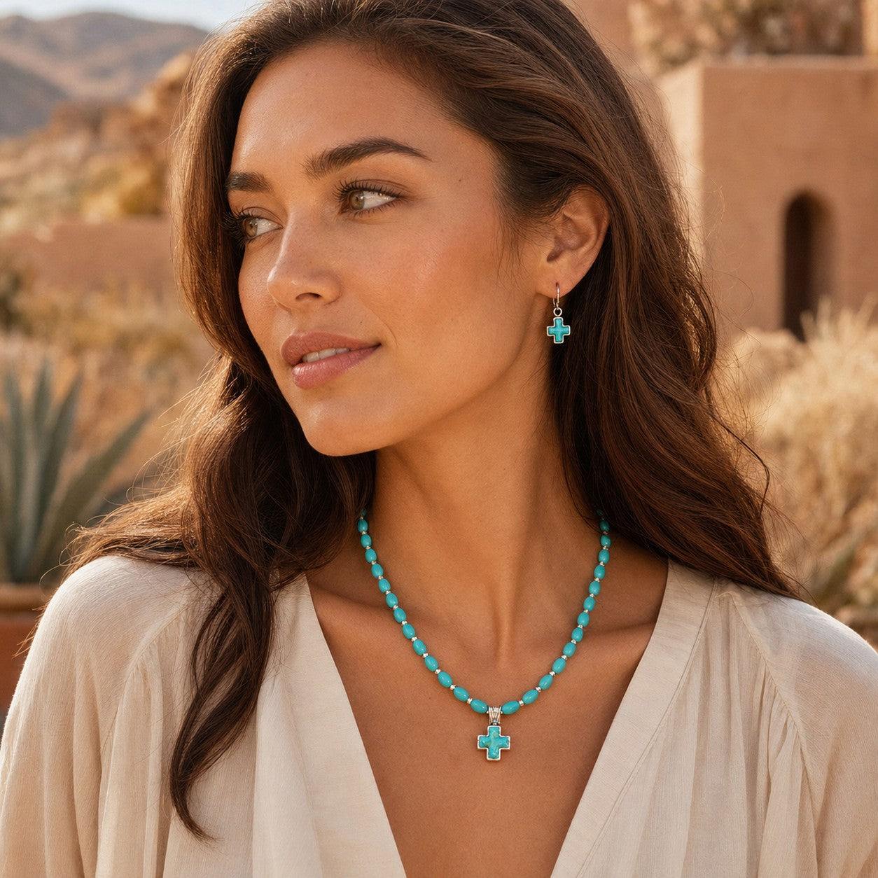 A woman wearing a turquoise beaded necklace with a cross pendant and matching drop earrings, photographed outdoors against a warm desert landscape.