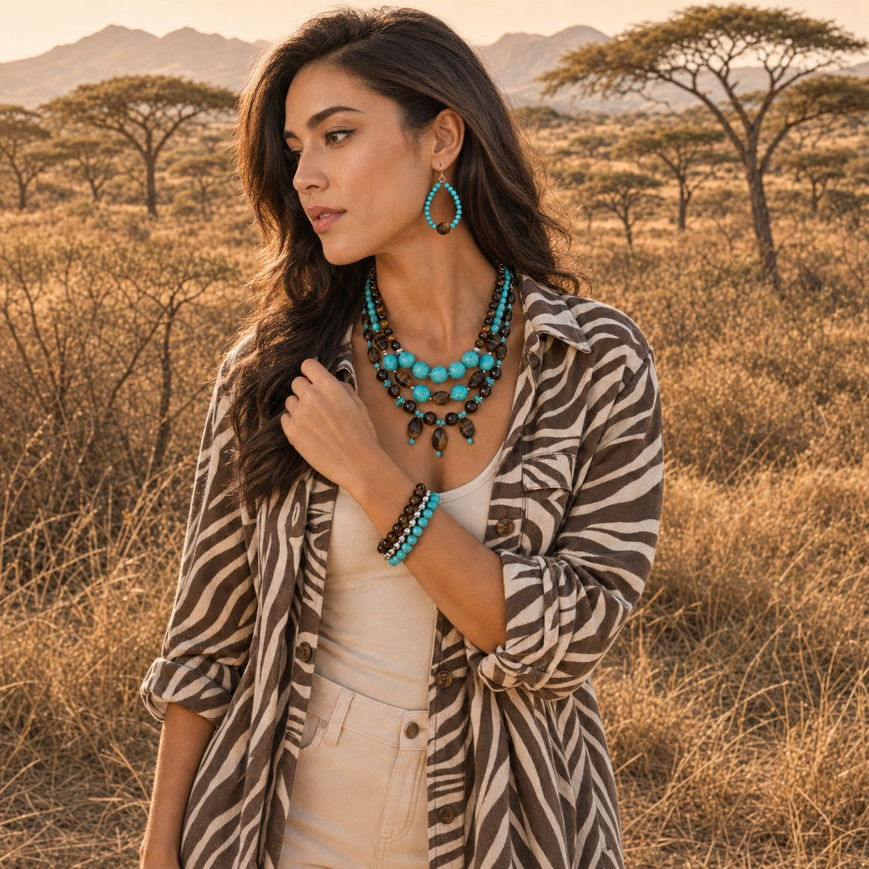 A woman stands in a dry grassy savanna landscape wearing a turquoise and black statement necklace, matching turquoise teardrop earrings, and a turquoise beaded bracelet, styled with a beige top and a brown-and-white zebra-print shirt.
