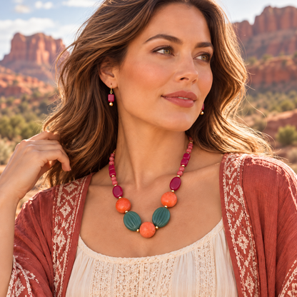 A woman wearing a colorful beaded necklace and matching drop earrings, photographed outdoors against a desert rock landscape. The necklace features large round beads in teal, orange, and magenta, paired with a matching beaded chain.