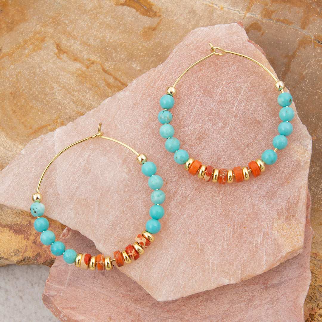 Gold hoop earrings with turquoise and orange beads on a stone surface
