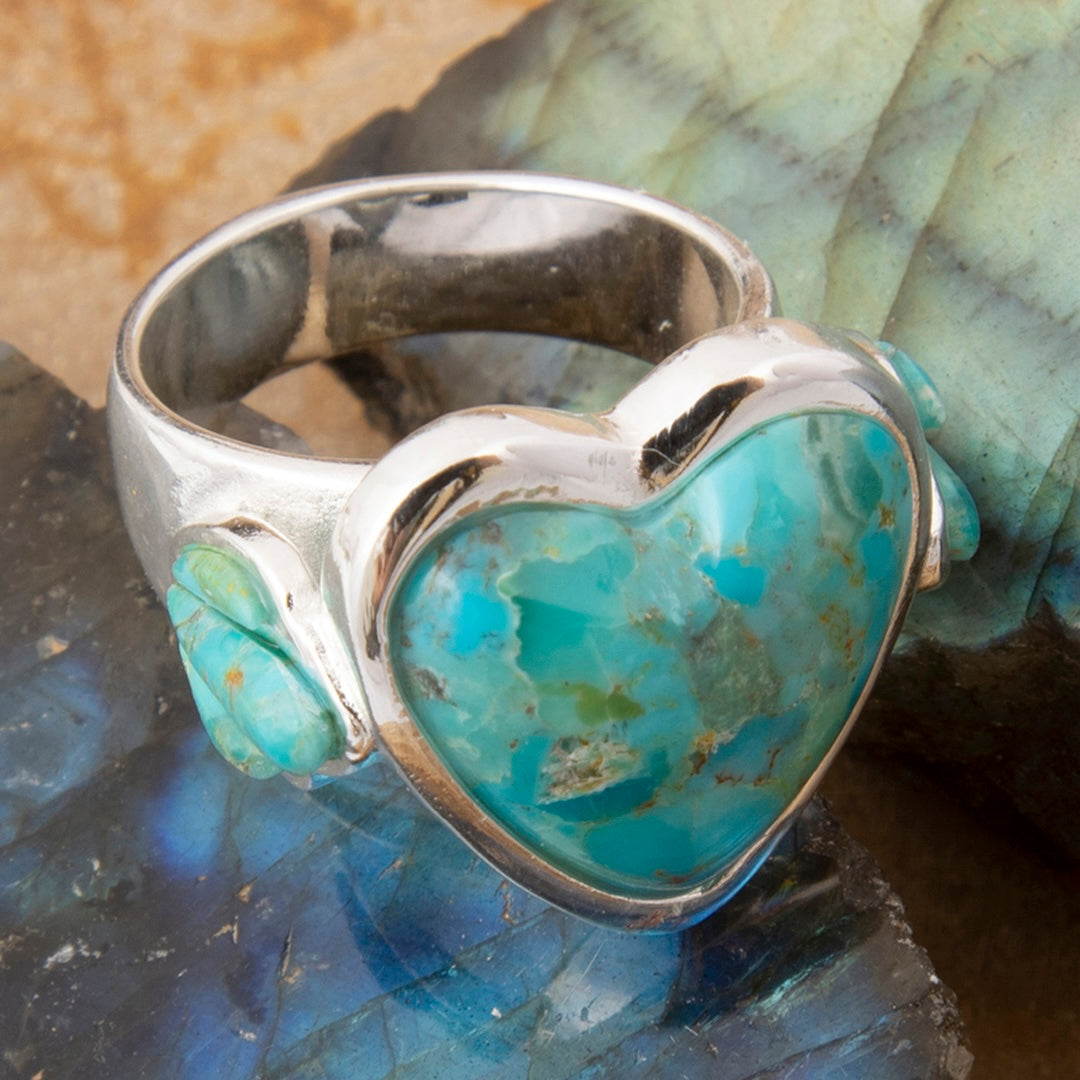 A sterling silver ring featuring a heart-shaped turquoise cabochon and a smaller matching turquoise stone on the side, displayed on a polished blue stone.