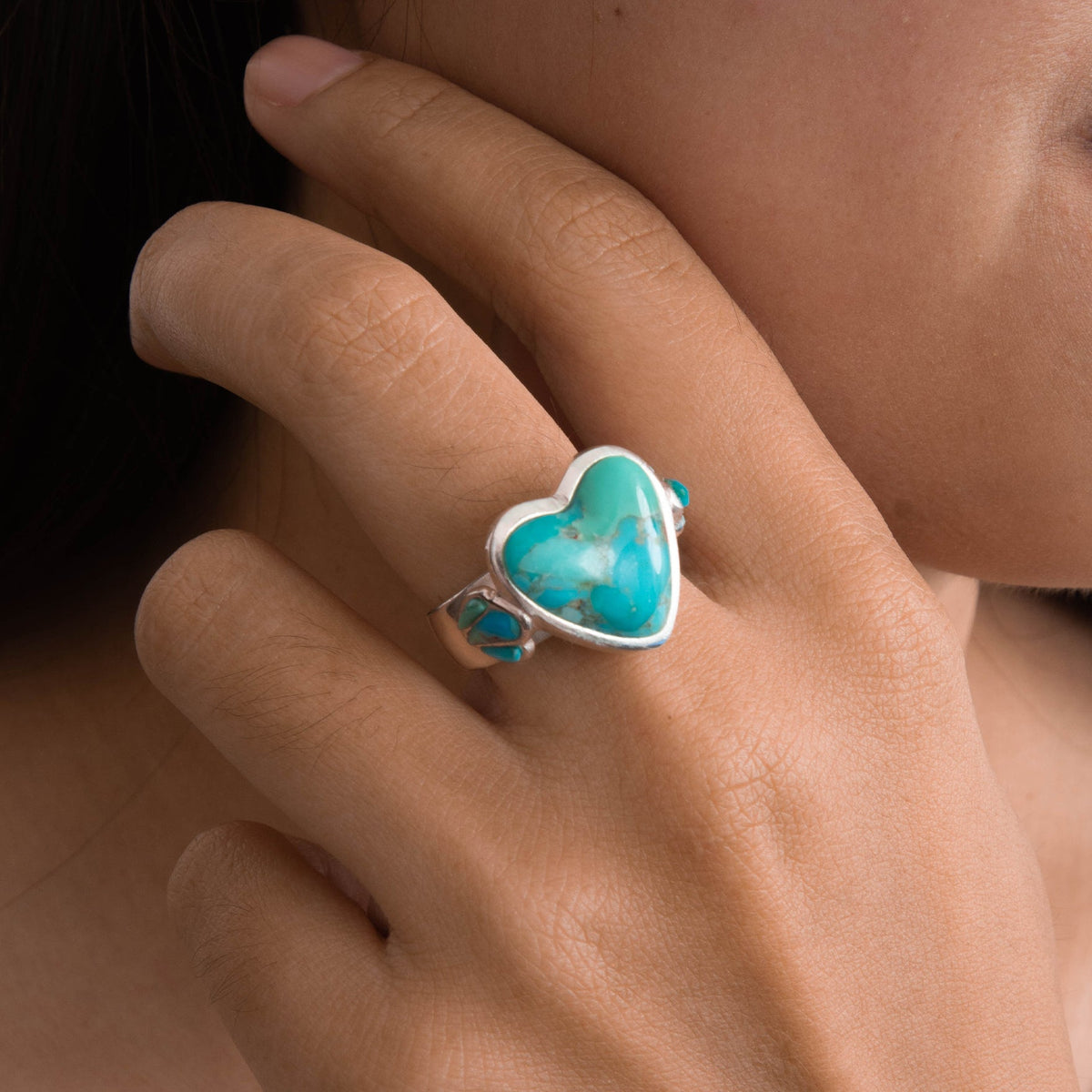 A close-up lifestyle shot of a person's hand wearing a silver ring with a heart-shaped turquoise cabochon and small round turquoise accents on the band.