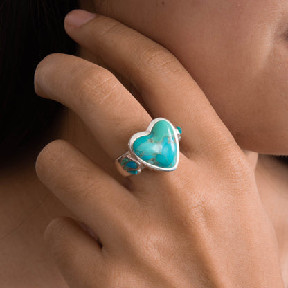 A close-up lifestyle shot of a person's hand wearing a silver ring with a heart-shaped turquoise cabochon and small round turquoise accents on the band.
