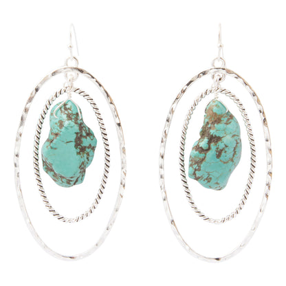 A pair of silver dangle earrings featuring organic-shaped turquoise stones with dark matrix, set within double textured oval hoops on a white background.