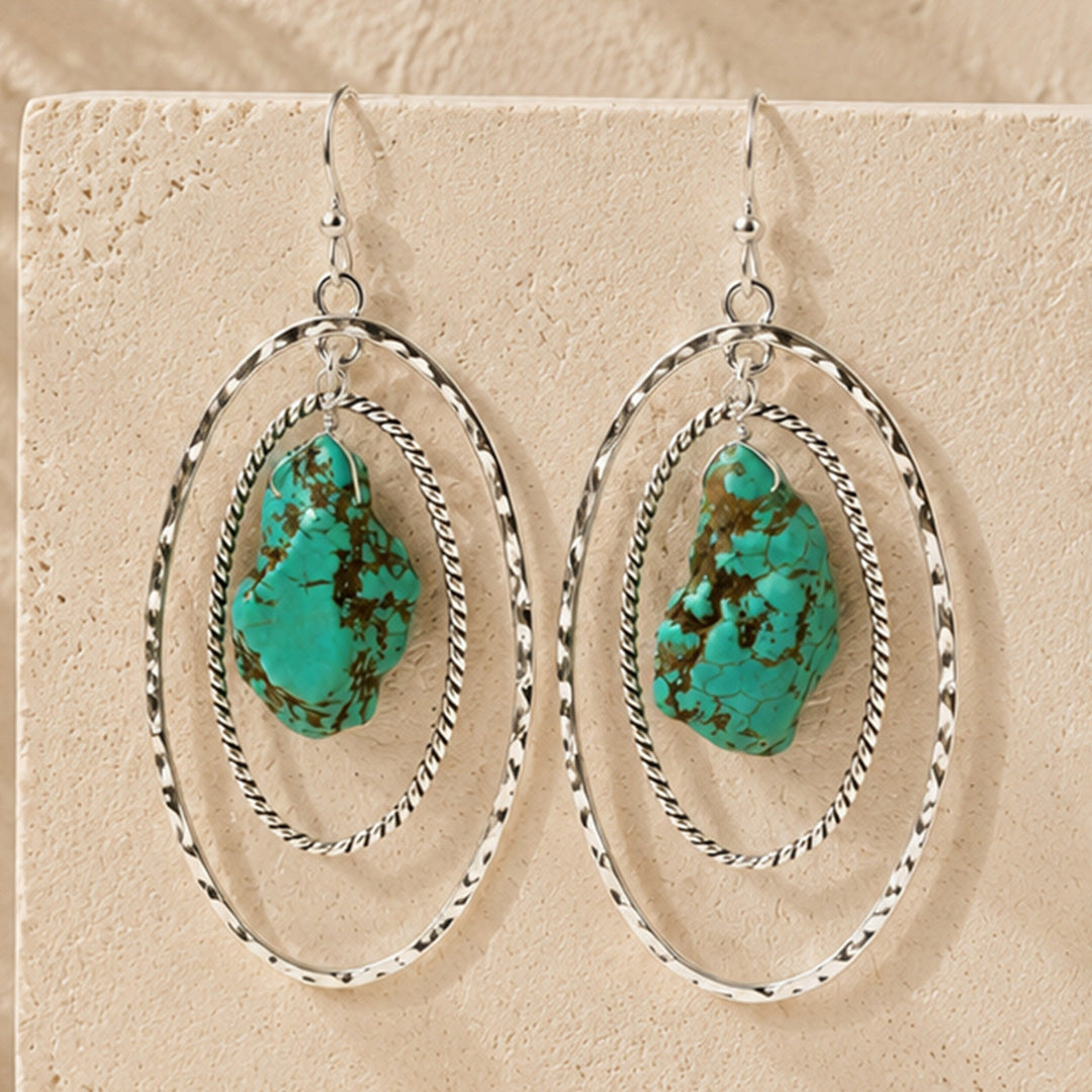 A pair of silver hoop dangle earrings with organic-shaped turquoise stones, displayed on a textured beige stone surface.
