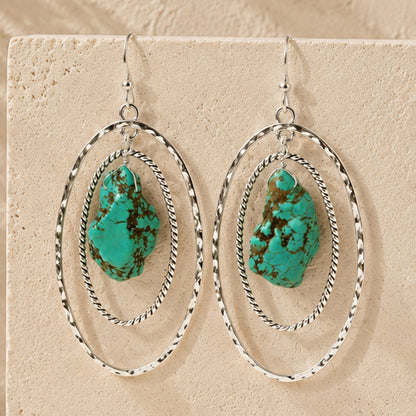 A pair of silver hoop dangle earrings with organic-shaped turquoise stones, displayed on a textured beige stone surface.