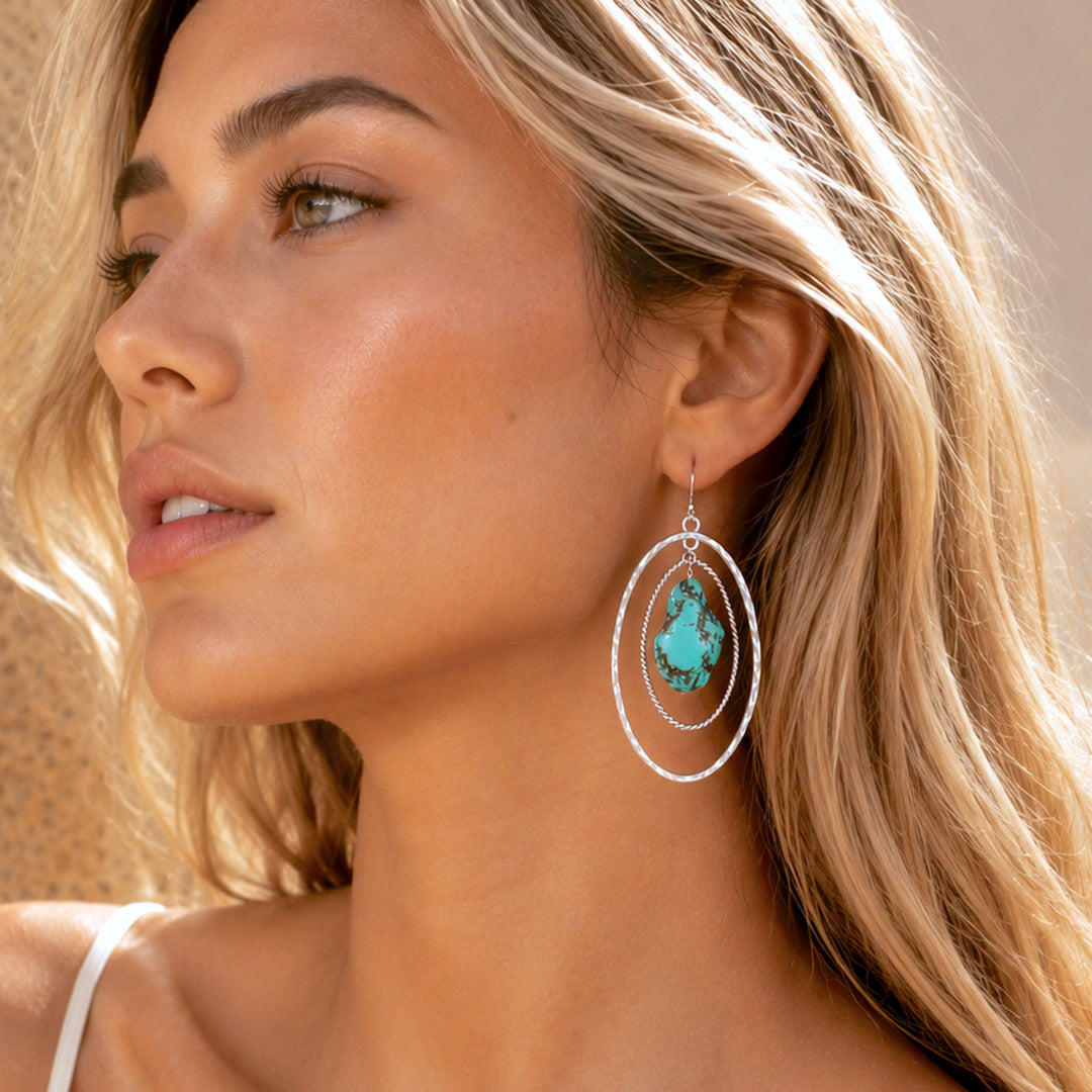 A close-up lifestyle shot of a woman wearing a large silver-toned dangle earring with a teardrop-shaped turquoise stone.