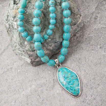 Turquoise beaded necklace with a large teardrop-shaped turquoise pendant in a silver bezel, draped over a gray rock on a textured gray surface.