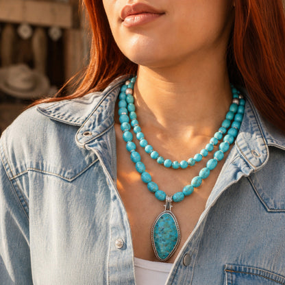 A close-up of a woman wearing a double-strand turquoise beaded necklace with a large oval turquoise pendant, styled with a light blue denim shirt.