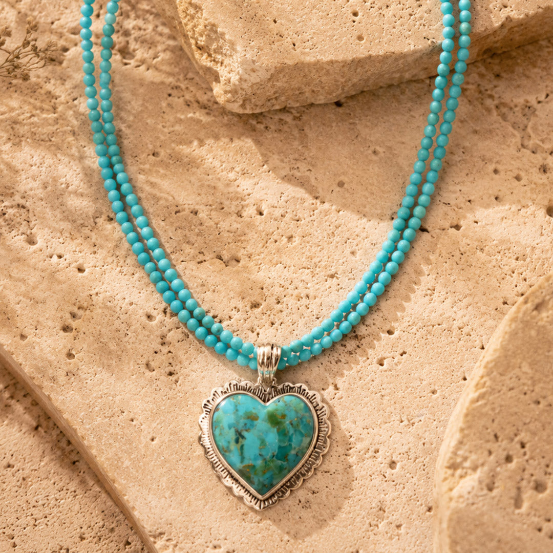 A turquoise heart pendant necklace with a silver-toned filigree border, hanging from a double-strand turquoise beaded necklace, displayed on textured tan stone.