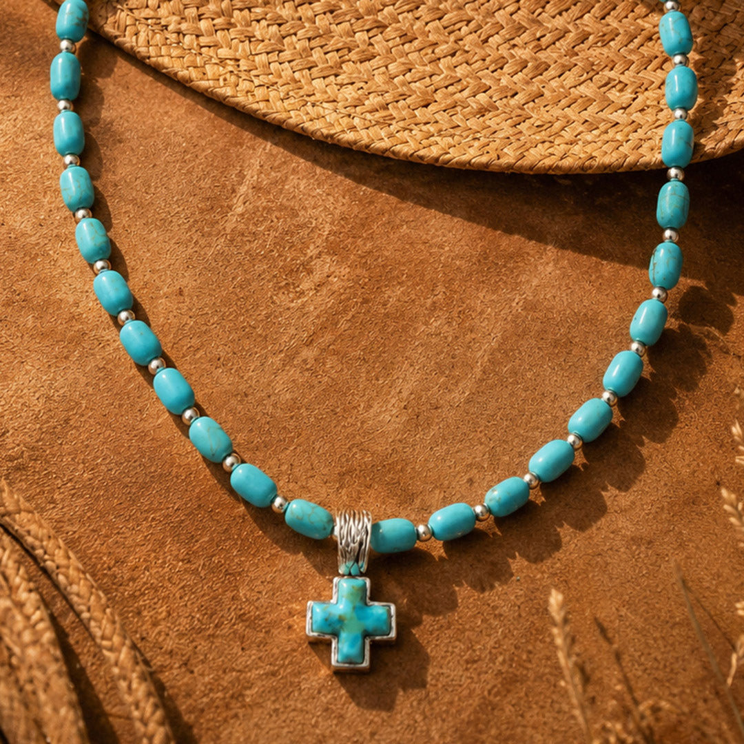 A turquoise and silver cross pendant necklace displayed on a textured brown surface next to a woven straw hat.