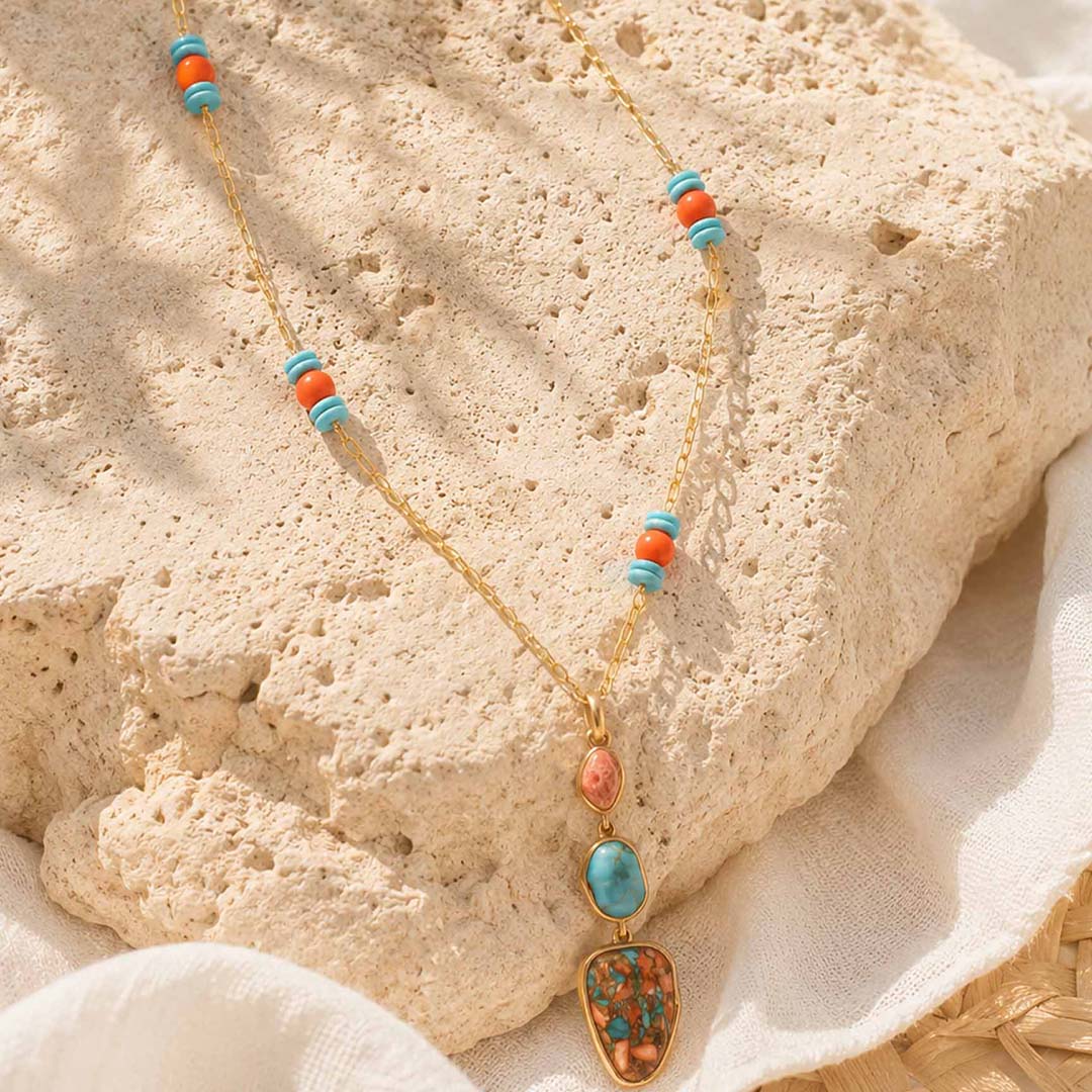 A gold chain necklace with turquoise and coral beads and a colorful stone pendant, styled on a textured beige stone block with white fabric.