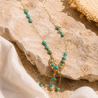 A gold-toned necklace with turquoise blue-green beads and a crescent moon pendant, styled on a beige stone with white fabric.
