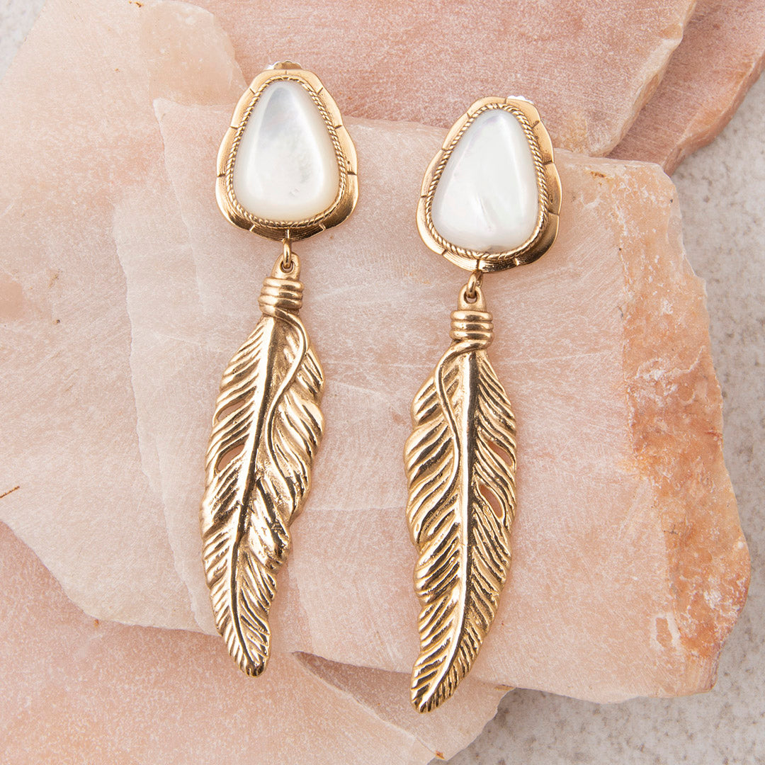 Gold feather earrings with white stones on a stone surface