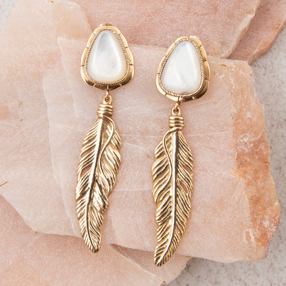 Gold feather earrings with white stones on a stone surface